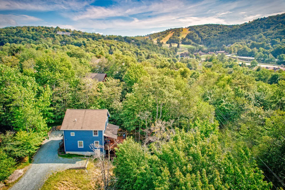 A wide view of the blue house with surrounding trees and distant hills. The porch is visible, inviting guests to start or end their day immersed in mountain scenery. The landscape hints at hiking trails and exploration opportunities just beyond the doorstep.