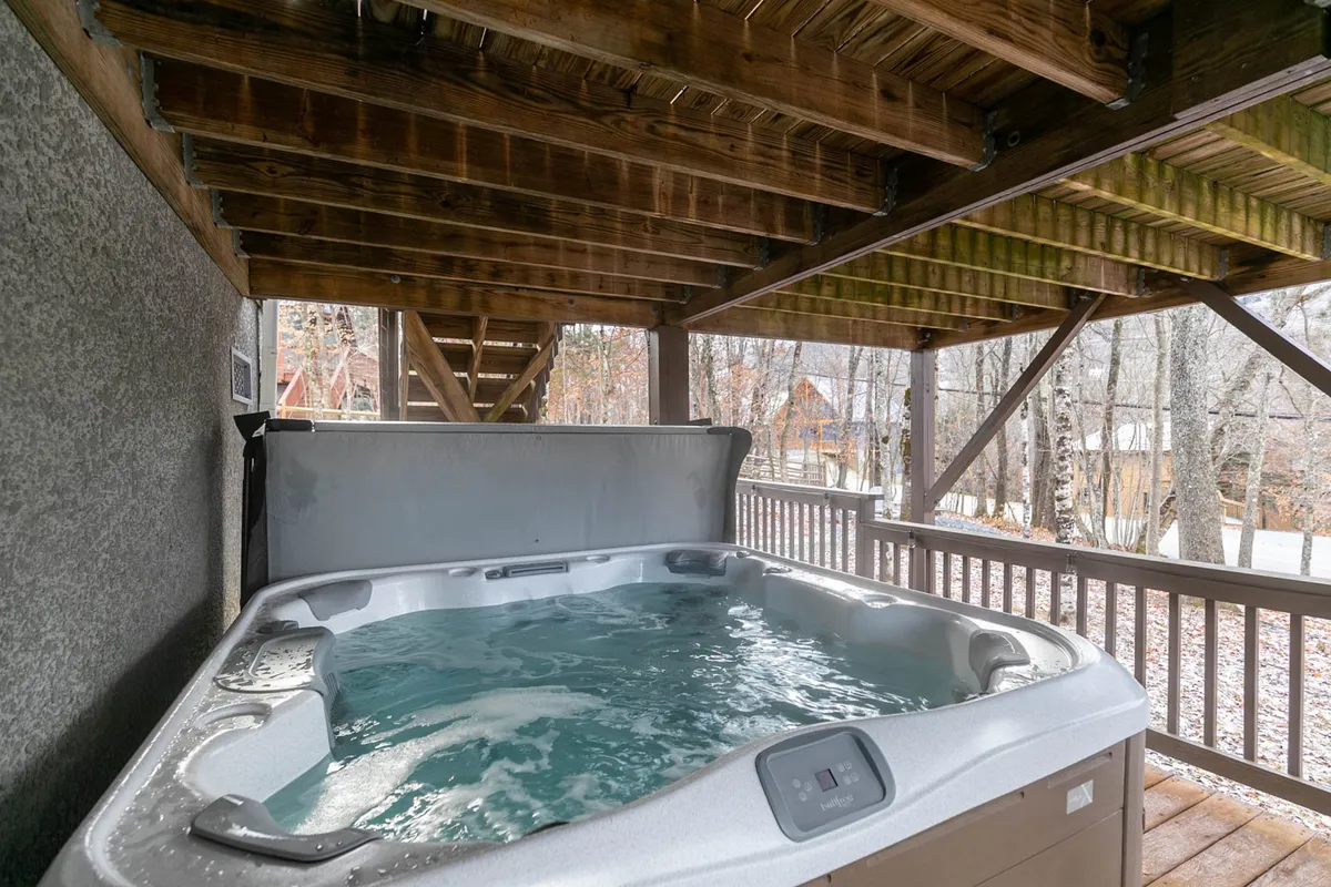 A covered deck area featuring a private hot tub, surrounded by wooden beams and leafy trees. The tranquil setting is ideal for year-round relaxation and soaking while taking in the peaceful mountain scenery.