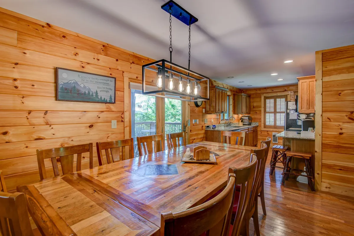 The image features a large wooden dining table with pendant lighting and surrounding windows that let in natural light. The space is designed for group meals, conversations, and games, perfectly suited for ten guests as described. The cozy wood paneling adds mountain lodge charm, and the visual emphasis is on the communal dining experience.