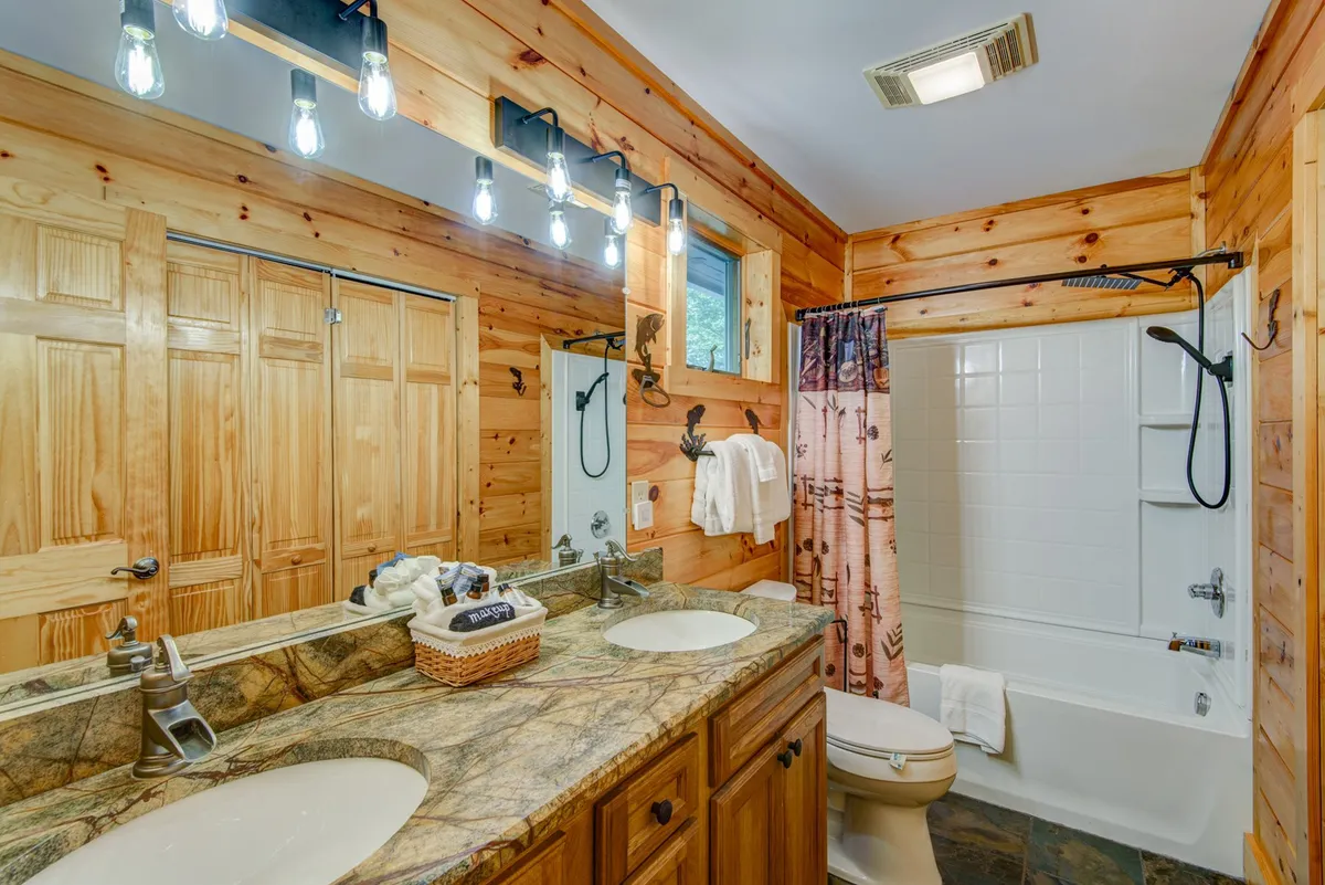 A well-appointed bathroom featuring a double sink granite vanity, wooden cabinetry, large mirrors, and a decorative shower curtain. The space combines function and luxury for guests.