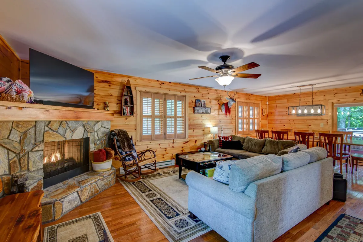 A cozy living area centers on a glowing fireplace, plush sofa, coffee table, and a large TV. The dining area is also visible, emphasizing the open flow between gathering and dining. Sunlight pours through shuttered windows, enhancing the inviting atmosphere. This is a true communal hub—ideal for relaxing, watching movies, or celebrating special occasions as mentioned in the section content.