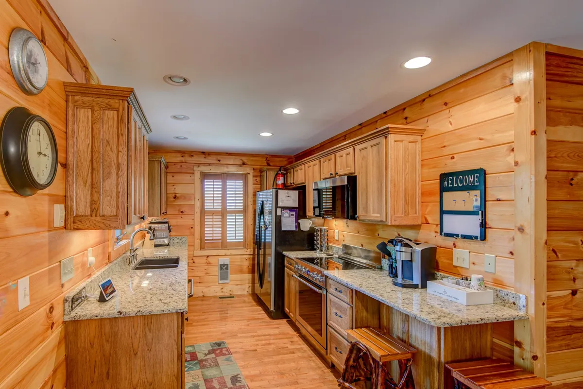 A close view of the kitchen area, featuring a granite countertop, wooden cabinets, modern appliances, and barstool seating. The 'WELCOME' sign and homey décor add warmth and character. The kitchen’s layout invites group cooking, coffee chats, and easy interaction with other parts of the main level.