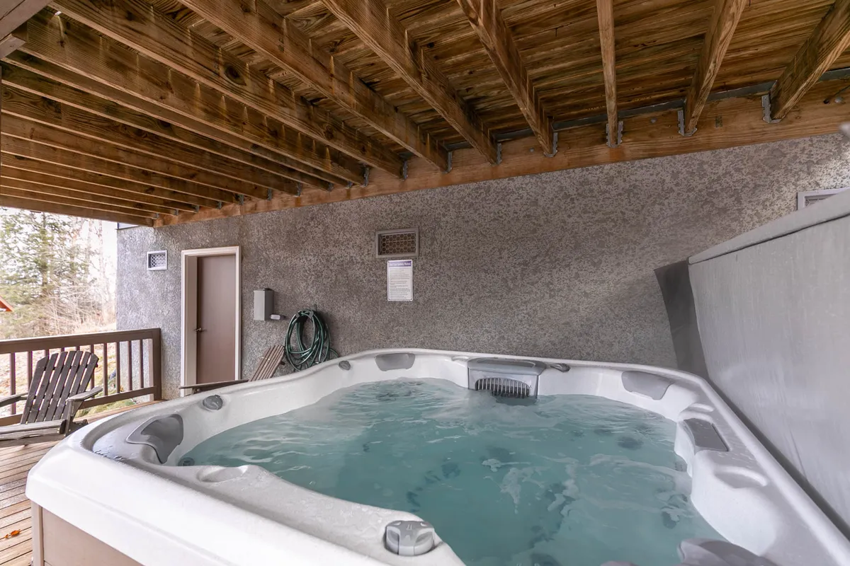 This image features a private hot tub set on a deck with rustic wooden beams and outdoor decking. The tub is positioned to allow for relaxation while enjoying views of the surrounding nature, with trees visible beyond the deck. The setup is inviting for groups or families to soak together while socializing or stargazing.