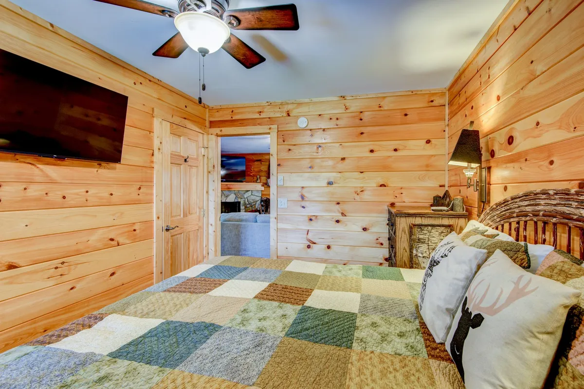 A tranquil bedroom with a large bed dressed in a plaid patchwork bedspread, rustic wooden walls, and cabin-style furniture. The ceiling fan and warm light create a welcoming retreat for restful nights.