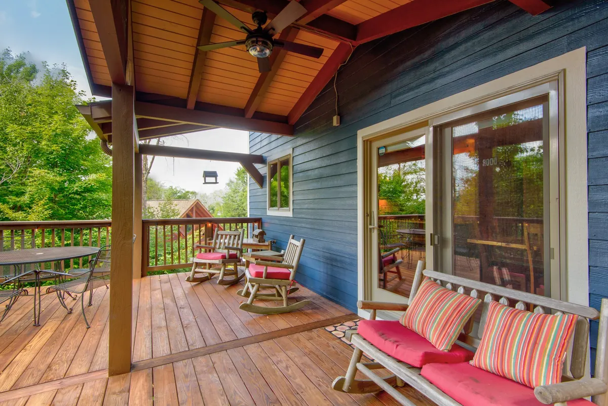 A cozy wooden deck featuring a table with chairs and inviting rocking chairs, all positioned next to a sliding glass door. The deck is surrounded by lush greenery, offering a seamless transition between indoor comforts and the natural mountain environment. The setup is ideal for relaxing while dogs can enjoy the secure outdoor space.