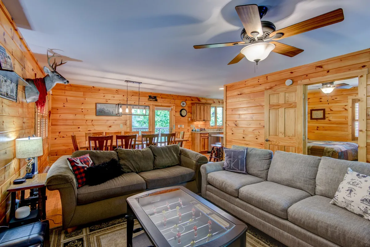This wide-angle view captures the heart of Dogwood Den’s main level, showing a comfortable seating area, a foosball table in the foreground, dining table for group meals, and the adjacent kitchen area. The space radiates warmth, with wood finishes and ample space for both relaxation and play. Multiple activity zones—game play, lounging, and dining—are visible and connected, highlighting the open-concept layout.