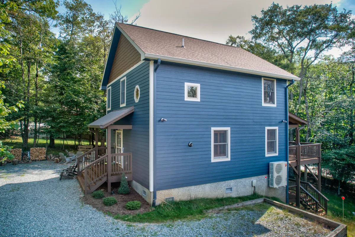 An exterior view of Dogwood Den showcasing its blue siding, expansive wooden deck, and lush surrounding trees. The deck is spacious, suitable for gatherings, and is depicted as an inviting spot for enjoying sunsets, nature walks, or outdoor meals.