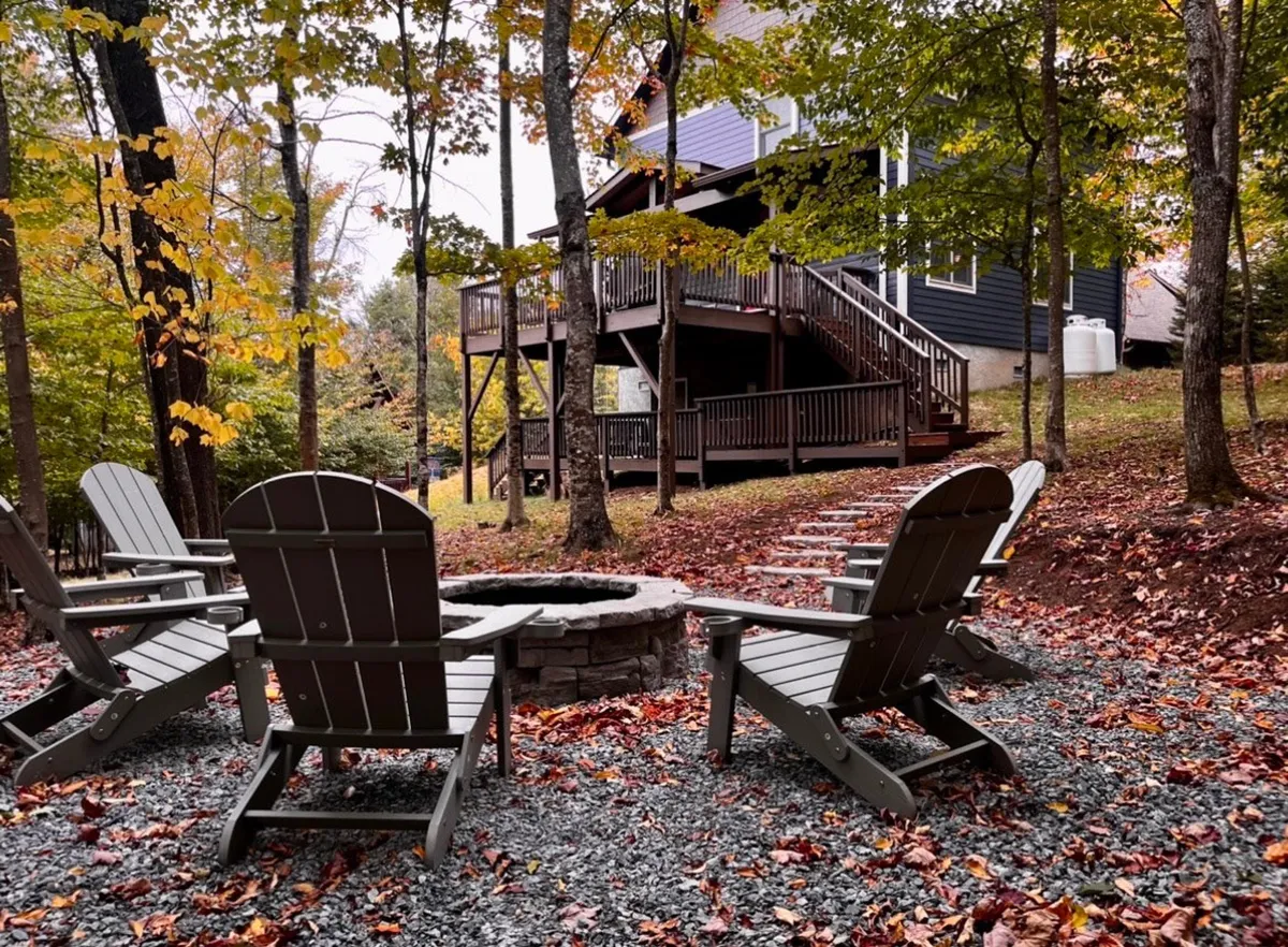 An inviting outdoor scene featuring Adirondack chairs encircled around a fire pit, set on the deck amidst tall trees. The mood is communal and relaxed, perfect for gathering after a day of hiking or skiing, with seasonal foliage visible in the background.
