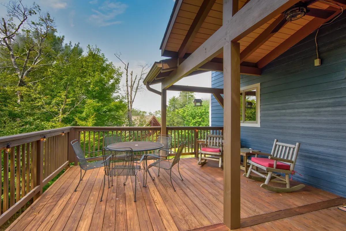 This image features a wooden deck with a circular table, rocking chairs, and lush green trees in the background. The calming setting invites guests to relax, dine outdoors, or simply enjoy the sights and sounds of nature right at their doorstep.